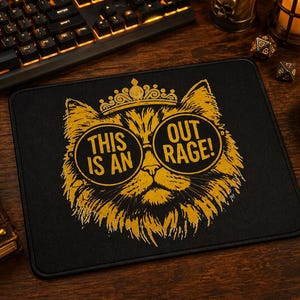 May include: Black rectangular mousepad featuring a gold illustration of a cat wearing glasses and a crown. The text on the mousepad reads "THIS IS AN OUTRAGE!". The mousepad is surrounded by a keyboard, mouse, and other desk accessories.