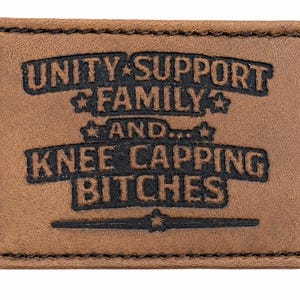 May include: A rectangular brown leather patch with black stitched edges. The patch features the words "UNITY SUPPORT FAMILY AND... KNEE CAPPING BITCHES" in a bold, stylized font. Stars and a horizontal line are also included.