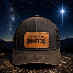 May include: A dark brown trucker hat featuring a rectangular tan leather patch on the front. The patch is embossed with the text "HE WHO FIGHTS WITH MONSTERS" in a bold, stylized font. The cap has a structured crown and a mesh back panel.