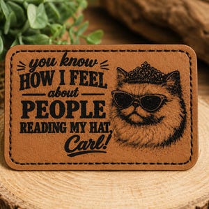 May include: A rectangular tan leather patch with black stitched edges. The patch features a laser-engraved illustration of a cat wearing a crown and sunglasses, alongside the text: you know HOW I FEEL about PEOPLE READING MY HAT, Carl!