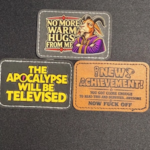 May include: Three rectangular morale patches with stitched edges. One features a goat in purple robes with the text NO MORE WARM HUGS FROM ME. Another is black with yellow text THE APOCALYPSE WILL BE TELEVISED. The third is tan with NEW ACHIEVEMENT text.