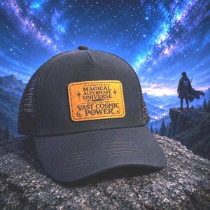 May include: A black trucker hat featuring a rectangular tan leather patch on the front. The patch is engraved with the text: I WENT TO A MAGICAL ALTERNATE UNIVERSE AND ALL I GOT WAS VAST COSMIC POWER, accompanied by small star and crescent moon icons.