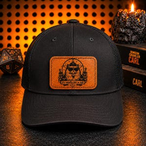May include: A black baseball cap with a mesh back and a rectangular tan leather patch on the front. The patch features a graphic of a cat wearing sunglasses inside a star, flanked by two candles, with the text "it's totally not a cult" below.