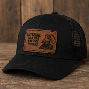 May include: A black baseball cap with a mesh back and a rectangular brown patch on the front. The patch features an engraved illustration of a goat with crossed arms and the text NO MORE WARM HUGS FROM ME in a bold, dark, sans-serif font.