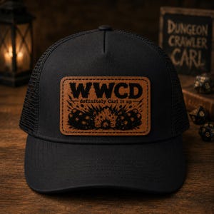 May include: A black trucker hat featuring a rectangular tan leather patch on the front. The patch displays the text WWCD definitely Carl it up above a graphic illustration of an explosion and two sticks of dynamite in black ink.