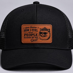 May include: A black trucker hat featuring a rectangular tan leather patch on the front. The patch displays a graphic of a cat wearing sunglasses and a crown, with the text: you know HOW I FEEL about PEOPLE READING MY HAT, Carl!