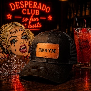May include: A black trucker hat featuring a rectangular tan leather patch on the front with the black text IWKYM. The hat has a structured front panel and a mesh back, positioned on a dark surface next to a silver tiara and a glass of red drink.