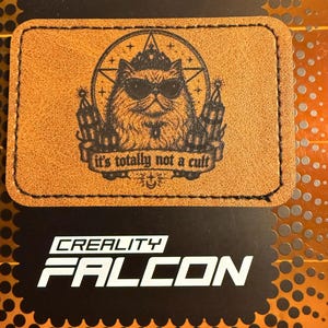 May include: A rectangular tan patch with black stitching around the edges. The center features a laser-engraved illustration of a cat wearing sunglasses and a crown inside a star, with the text "it's totally not a cult" on a banner below.