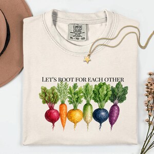 Peut inclure: Un t-shirt couleur crème avec une étiquette Comfort Colors en taille XL. Le t-shirt présente un graphique de six légumes racines colorés, dont une betterave rouge, une carotte orange, une betterave jaune, un radis blanc, une betterave bleue et une betterave violette avec le texte LET'S ROOT FOR EACH OTHER.