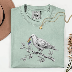 May include: A light green t-shirt featuring a black and white illustration of a dove perched on an olive branch, holding a small rainbow flag in its beak. The shirt has a Comfort Colors label at the neck, size XL, and is made of 100% cotton.
