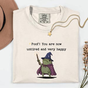 Può includere: Una t-shirt color crema con la grafica di una rana verde che indossa un cappello da mago blu e un mantello viola, tenendo in mano una bacchetta. Il testo recita: Poof! You are now untired and very happy. Etichetta Comfort Colors sul colletto.
