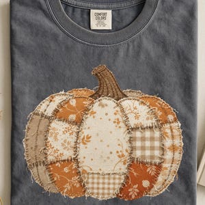 Può includere: Una t-shirt girocollo grigio antracite con una grande stampa di una zucca al centro. La zucca è composta da vari ritagli di tessuto nei toni dell'arancione, beige e bianco, con motivi floreali e a quadretti e bordi sfrangiati.
