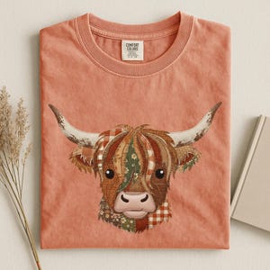 Op de afbeelding: Een terracotta-kleurig t-shirt met ronde hals en een afbeelding van een Schotse Hooglander-kop. De koe is samengesteld uit diverse patchwork-patronen in bruin-, oranje- en groentinten. Het shirt heeft een Comfort Colors merklabel in de nek.