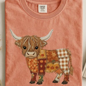 May include: A terracotta-coloured crew neck t-shirt featuring a central embroidered graphic of a Highland cow. The cow's body is composed of various patchwork patterns including autumn leaves, pumpkins, acorns, and a brown and white checkered print.