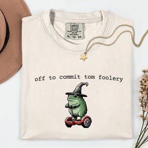 May include: A cream-coloured t-shirt featuring a graphic of a green frog wearing a black witch hat and riding a red self-balancing scooter. The text "off to commit tom foolery" is printed in black above the frog illustration on the front.