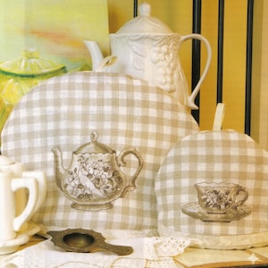 Puede incluir: Naturaleza muerta con una tetera de cerámica blanca con diseño de uvas, un cubre tetera y un cubre taza. Los cubre tetera y taza son a cuadros beige y blancos. También se ve un colador de té vintage.