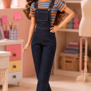 May include: A doll with long brown hair wearing a pink cap, an orange and blue striped short-sleeve shirt, dark blue denim overalls with metal buttons, and white sneakers with orange side stripes. The doll has a fair complexion and blue eyes.