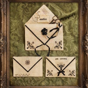May include: Framed artwork featuring three linen pouches with black embroidered details and accents. The top pouch holds scissors and a label that reads "needles." The bottom pouches have text that reads "still stitching" and "created in the year..."