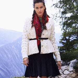 May include: A woman wearing a cream-colored cable-knit cardigan with a black belt and a black skirt. A red patterned scarf is worn under the cardigan. The background features a mountain range and trees.