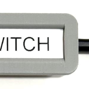 Peut inclure: Un support d'étiquette gris avec le mot "SWITCH" imprimé sur une étiquette blanche, attaché à un câble réseau noir. Le câble a un connecteur RJ45 transparent sur le côté droit.