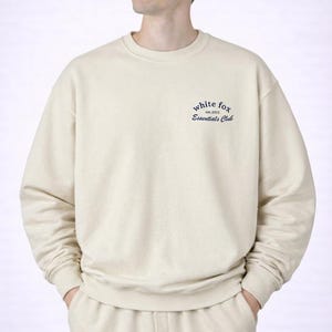 Peut inclure: Ensemble de survêtement crème composé d'un sweat-shirt et d'un pantalon assorti. Le sweat-shirt a un col rond et le texte "white fox est. 2013 Essentials Club" en bleu marine sur la poitrine gauche. Le modèle porte l'ensemble.