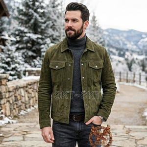 May include: An olive green suede jacket with button fastenings and chest pockets. The jacket is worn with a grey turtleneck and dark wash jeans. The background features a snowy mountain landscape.