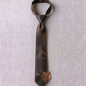 May include: A brown leather tie with a textured finish. The tie is knotted at the top and has a pointed bottom. A circular emblem with an eagle design is near the bottom. The tie is displayed against a neutral background.