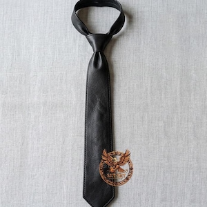 May include: A black leather tie with a pointed end, knotted at the top. The tie is displayed on a light gray textured surface. A circular emblem with a brown eagle is at the bottom of the tie.