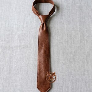 May include: A brown leather tie, knotted at the neck, lies against a neutral background. The tie is a rich, warm brown color, with visible texture and stitching along the edges. The tie's design is classic and elegant, suitable for formal or business attire.