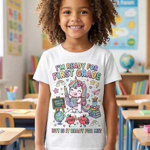 Könnte beinhalten: Ein weißes T-Shirt mit einer bunten Grafik eines Einhorns, das ein Buch liest, umgeben von Schulsachen wie Äpfeln, einem Globus und Büchern. Der Text lautet I'M READY FOR FIRST GRADE BUT IS IT READY FOR ME? in mehrfarbigen Buchstaben.