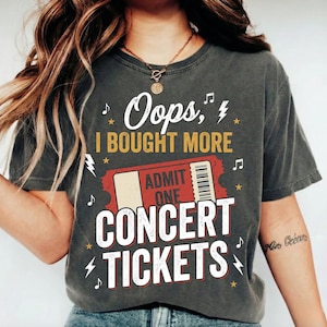 以下が含まれることがあります： 「Oops, I BOUGHT MORE CONCERT TICKETS」という白と黄色の文字と、赤い「ADMIT ONE」チケットのグラフィックがプリントされたチャコールグレーの半袖Tシャツ。デザインには小さな白い音符、稲妻、黄色い星が含まれています。