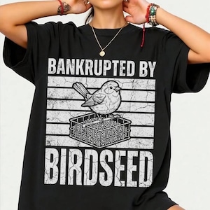Könnte beinhalten: Ein schwarzes Kurzarm-T-Shirt mit einem weißen Grafikdruck eines Vogels, der auf einem Vogelfutterspender sitzt. Der Text "BANKRUPTED BY" steht über dem Vogel und "BIRDSEED" unter dem Futterspender, alles vor einem abgenutzten Hintergrund.