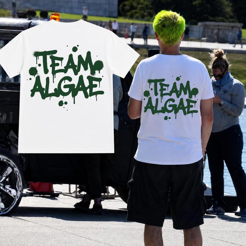 Puede incluir: Una camiseta blanca de manga corta con el texto "TEAM ALGAE" impreso en una fuente de estilo grafiti de color verde oscuro con efectos de goteo de pintura. El dise&ntilde;o est&aacute; centrado en la parte posterior de la camiseta.