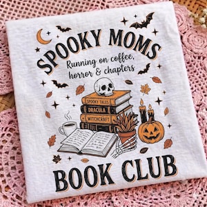 Könnte beinhalten: Ein weißes T-Shirt mit einem Grafikdruck und dem Text "SPOOKY MOMS Running on coffee, horror & chapters BOOK CLUB". Das Design zeigt einen Bücherstapel, einen Totenkopf, eine Kaffeetasse, eine Topfpflanze, einen Kürbis und Fledermäuse.