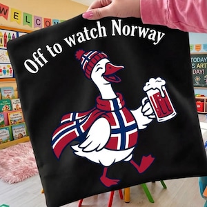 Könnte beinhalten: Eine schwarze Tragetasche mit einer weißen Grafik einer Gans, die eine Mütze und einen Schal im Muster der norwegischen Flagge trägt. Die Gans hält einen Bierkrug mit Schaum. Der Text "Off to watch Norway" ist in Weiß darüber gedruckt.