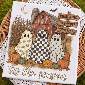 Könnte beinhalten: Ein weißes quadratisches Schild mit drei Geistern mit Gänseblümchen-, Schachbrett- und Punktemustern vor einer roten Scheune, Maisfeldern und Kürbissen. Ein Holzschild zeigt PUMPKINS, HAY-RIDES, CORN MAZE. Unten steht der Text 'tis the season'.