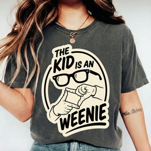 Könnte beinhalten: Ein anthrazitfarbenes Kurzarm-T-Shirt mit einem cremefarbenen Grafikdesign und schwarzem Text, der THE KID IS AN WEENIE lautet. Die Grafik zeigt eine schwarz umrandete Brille und zwei Hände, die einen quadratischen Rahmen bilden.