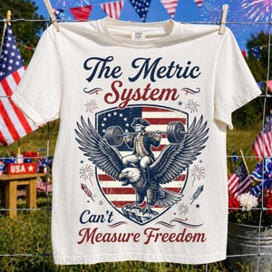 Könnte beinhalten: Ein cremefarbenes Kurzarm-T-Shirt mit einer Grafik eines Weißkopfseeadlers, der eine Person in Kolonialkleidung trägt, die eine Langhantel hebt. Der Text lautet The Metric System Can't Measure Freedom, umgeben von Feuerwerk.