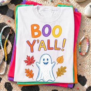 Könnte beinhalten: Ein weißes T-Shirt mit dem Text "BOO Y'ALL!" in orangefarbenen, violetten und gelben Buchstaben. Unter dem Text befindet sich eine weiße Geisterillustration, umgeben von roten, orangefarbenen und gelben Herbstblättern. Das T-Shirt liegt auf anderen.