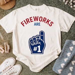 Könnte beinhalten: Ein weißes Kurzarm-T-Shirt mit dem roten Schriftzug FIREWORKS, dem blauen Schriftzug ARE und einer großen blauen Schaumstofffinger-Grafik mit dem weißen Text #1 darunter. Das Design ist auf der Vorderseite des Baumwollshirts aufgedruckt.