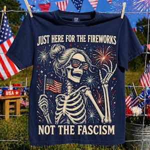 Könnte beinhalten: Ein marineblaues T-Shirt mit einer Grafik eines Skeletts mit Haarknoten, das eine Sonnenbrille im US-Flaggen-Design trägt. Das Skelett hält eine kleine US-Flagge. Feuerwerk umgibt die Figur. Der Text lautet: JUST HERE FOR THE FIREWORKS NOT THE FASCISM.