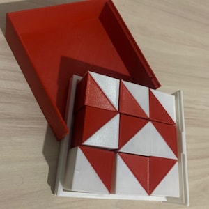 以下が含まれることがあります： 赤い蓋が付いた赤と白のパズルキューブ、白いトレイに入っています。 キューブは、幾何学模様を作成する小さな三角形のブロックで構成されています。 パズルは、操作と問題解決のために設計されています。