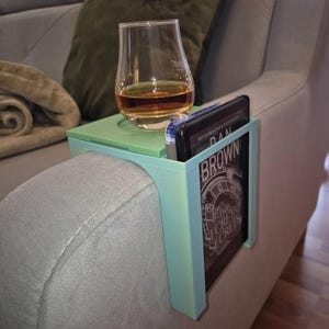 May include: A mint green and light green sofa arm table with a built-in drink holder and a pocket for a tablet or book. A glass of amber liquid sits in the drink holder. The book in the pocket has the author's name, Dan Brown, visible.