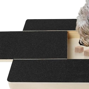 Op de afbeelding: Een hondenpuzzelspeelgoed met een zwart oppervlak en een schuifcompartiment. Het compartiment bevat verschillende kleine, vierkante traktaties. Een hand trekt aan een lipje om het schuifcompartiment te openen. Een hond eet de traktaties.