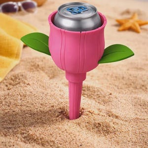 May include: A pink tulip-shaped drinks holder with green leaves, holding a silver can. The holder is stuck in the sand on a beach, with a beach bag, sunglasses, and a starfish in the background. A yellow towel is also visible.