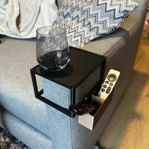 May include: A black, square armrest table with a glass of water, a remote control, and a media remote holder. The table is attached to a gray couch with a patterned pillow and a blanket.