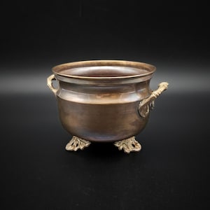 May include: A bronze-colored metal cauldron with ornate handles and feet. The cauldron has a rounded body and a wide opening. The handles and feet feature detailed, decorative designs. The cauldron is set against a black background.