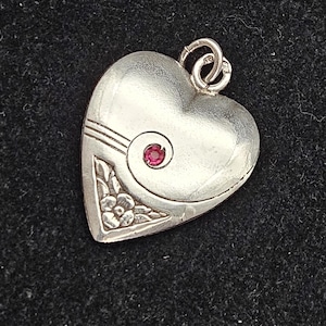 May include: A silver-toned heart-shaped pendant featuring a small, circular red gemstone set within a spiral design. The lower left corner displays an engraved floral pattern with leaves and a flower, complemented by three horizontal lines.