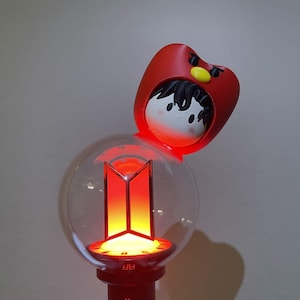 Op de afbeelding: Een lichtstaaf met een doorzichtige bolvormige bovenkant met een gloeiend oranje en rood BTS-logo. Een rode handgreep ondersteunt de bol. Een figuurtje met zwart haar en een rode capuchon met een gele snavel en zwarte wenkbrauwen zit bovenop.