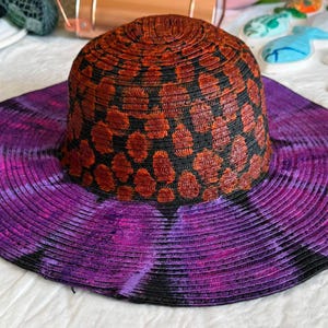 May include: A wide-brimmed straw hat featuring a crown with a black and burnt orange leopard print pattern. The wide brim displays a vibrant tie-dye design in shades of deep purple and black, creating a distinct contrast with the patterned crown.